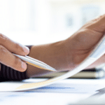 This image depicts a close-up of someone's hands holding a document, with one hand pointing at it using a pen. The scene appears to take place in a professional or office setting, with papers and possibly graphs or data visible on the desk. The natural lighting and composition emphasize focus and productivity.