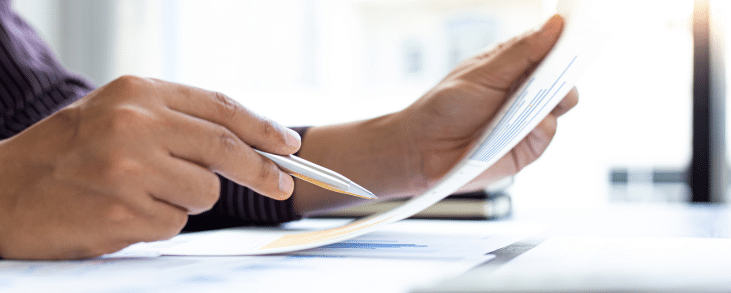 This image depicts a close-up of someone's hands holding a document, with one hand pointing at it using a pen. The scene appears to take place in a professional or office setting, with papers and possibly graphs or data visible on the desk. The natural lighting and composition emphasize focus and productivity.