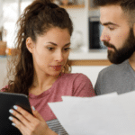 Couple sitting on a couch in a bright, modern living room. The woman, holding a tablet, looks at some documents that the man is holding, suggesting they are reviewing or discussing something important, possibly financial or household-related. The setting appears casual yet focused