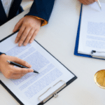 The image shows a professional setting where two individuals are working on documents. One person is writing on a clipboard, while another is holding a pen over a separate clipboard. The focus is on their hands and the documents. On the desk are additional items, including the scales of justice, suggesting a legal or contractual context, such as signing agreements or legal paperwork.