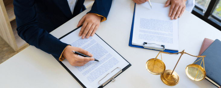 The image shows a professional setting where two individuals are working on documents. One person is writing on a clipboard, while another is holding a pen over a separate clipboard. The focus is on their hands and the documents. On the desk are additional items, including the scales of justice, suggesting a legal or contractual context, such as signing agreements or legal paperwork.