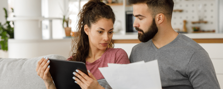 Couple sitting on a couch in a bright, modern living room. The woman, holding a tablet, looks at some documents that the man is holding, suggesting they are reviewing or discussing something important, possibly financial or household-related. The setting appears casual yet focused